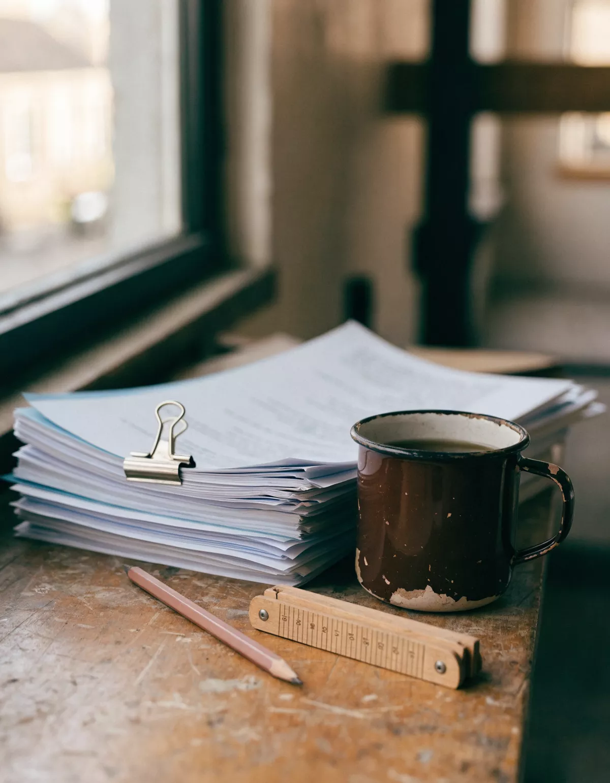 A thick printed document pack held with a bulldog clip on a workbench, beside an enamel mug, a carpenter's pencil and a folding rule.