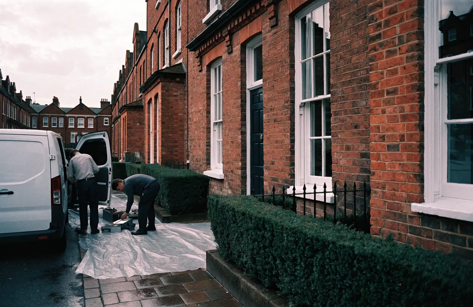 Two tradespeople unloading tools from a van onto a dust-sheeted path outside a British red-brick terraced house.